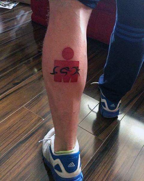 Tattoo of the Ironman logo in red with unique detail, featuring a personal touch with stylized numbers beneath, symbolizing the athlete's personal race or achievement.