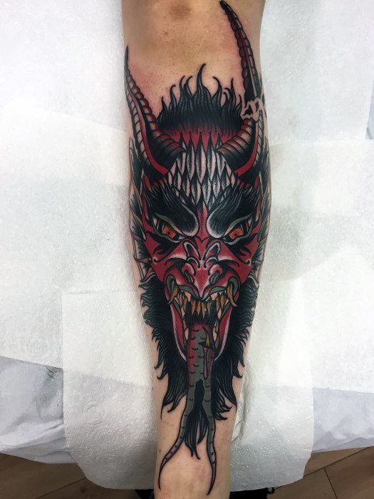 Striking Krampus tattoo on the leg featuring a fierce red and black horned face, sharp fangs, glowing eyes, and a forked tongue.