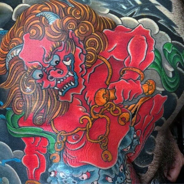 Bright Raijin chest tattoo with red thunder god, orange chains and drumsticks, flowing hair, and swirling green and blue clouds.