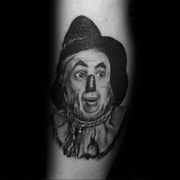 Realistic black and grey tattoo of the classic Scarecrow character from *The Wizard of Oz*, featuring expressive face and rope collar.