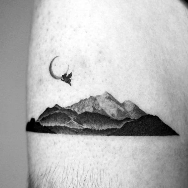 Minimalist mountain tattoo on the arm, featuring soft shading, a crescent moon, and a floating figure for a dreamy, surreal landscape design.