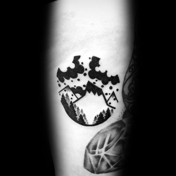 Bold circular mountain tattoo on the arm, featuring black ink clouds, trees, and geometric peaks for a striking and modern nature-inspired design.