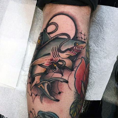 Neo-traditional shark tattoo entangled in an anchor and rope, featuring fierce expression, dark shading, and pops of red and yellow.