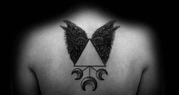 Back tattoo of Huginn and Muninn facing outward with wings forming a triangle, combined with crescent moons and geometric symbols.