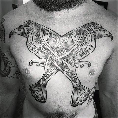 Nordic-style chest tattoo of Huginn and Muninn as mirrored axe-headed ravens with runes and intricate Viking patterns across the pectorals.