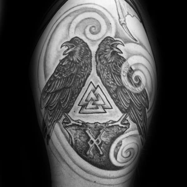 Black and grey upper arm tattoo of Huginn and Muninn flanking the Valknut, with swirling background and Norse rune details.