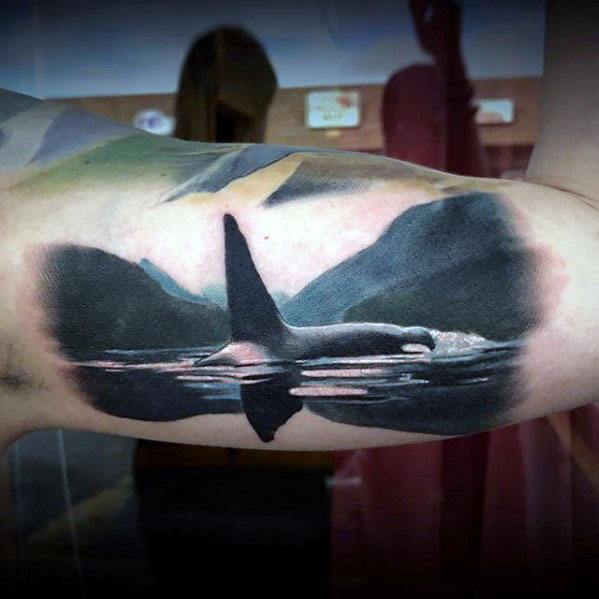 Realistic black and gray orca tattoo on the forearm, showcasing the whale's dorsal fin above the water, with a scenic backdrop for added depth.