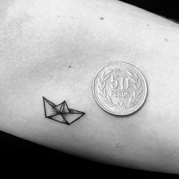 Tiny minimalist paper boat tattoo next to a 50 pesos coin, symbolizing simplicity, nostalgia, and life’s journey in a subtle design.
