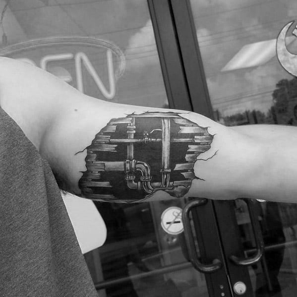Realistic black and grey tattoo on the inner bicep showing plumbing pipes beneath torn skin, revealing a hidden mechanical core.