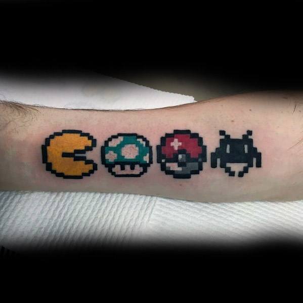 Retro gaming tattoo featuring 8-bit icons of Pac-Man, Super Mario mushroom, Pokeball, and Space Invader lined up on the forearm.