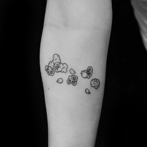 Minimalist black ink tattoo of scattered popcorn kernels and popped corn pieces across the inner forearm in a simple, clean style.