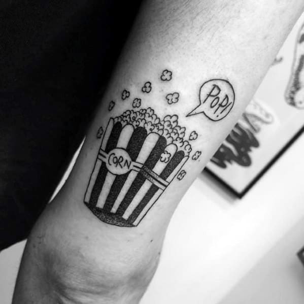 Blackwork tattoo of a striped popcorn bucket with the word CORN and a speech bubble saying POP, with small kernels floating around.