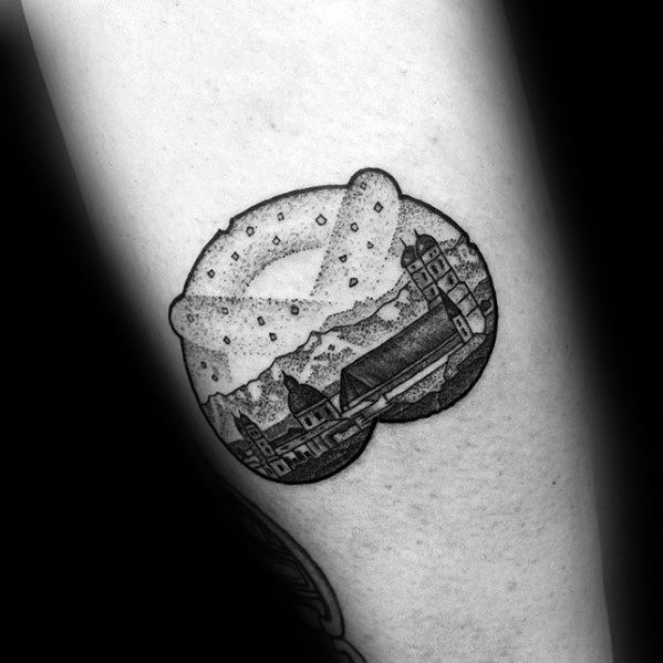 Black and grey pretzel tattoo with intricate dotwork, featuring a scenic village and mountain landscape inside the lower half of the design.