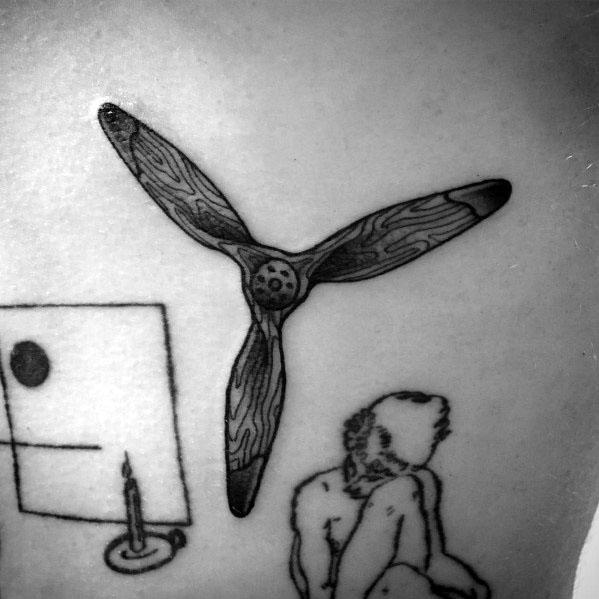 Black and grey tattoo of a three-blade wooden propeller with fine line detailing, placed among abstract and artistic elements on the skin.