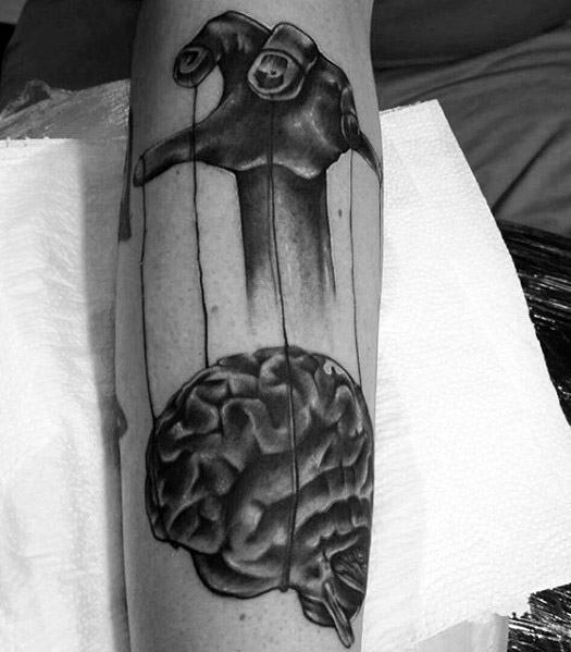 Black and grey puppet tattoo of a hand controlling a brain with strings, symbolizing mental manipulation or control over thoughts.
