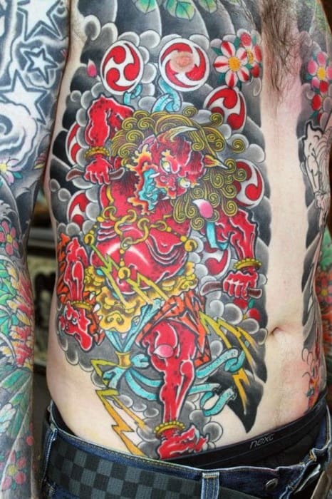 Colorful Raijin torso tattoo with vivid red thunder god, swirling clouds, yellow lightning bolts, and floral patterns in bold detail.