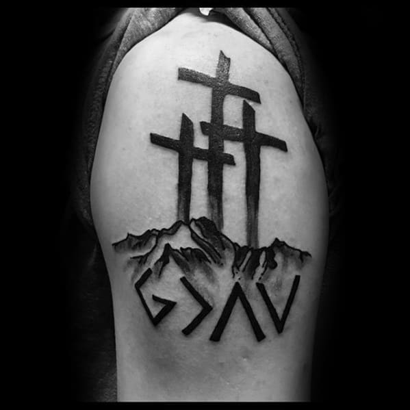 Upper-arm tattoo integrates the "God is greater than the highs and lows" symbol with a bold depiction of three crosses atop a mountainous landscape, emphasizing faith, sacrifice, and spiritual strength