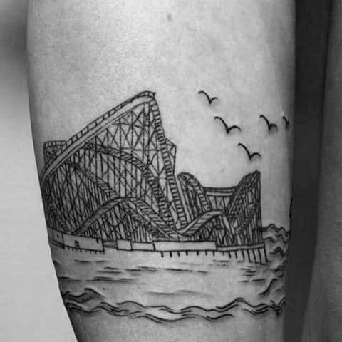 Linework roller coaster tattoo beside ocean waves, with wooden structure detail and birds flying overhead.
