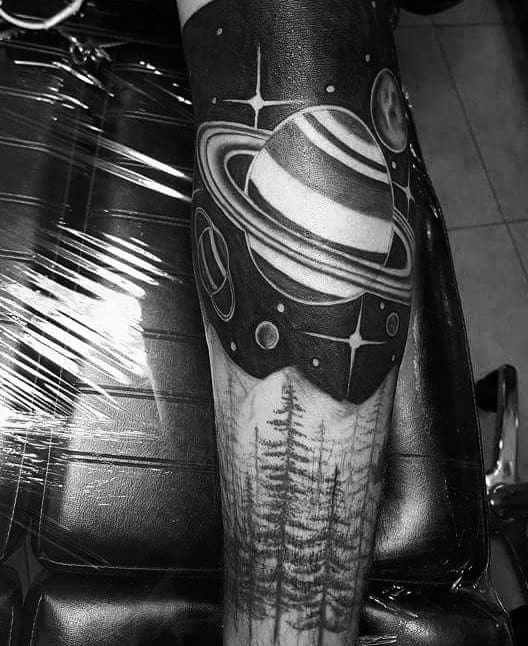 Black and grey Saturn tattoo design for men combining outer space with forest trees, planets, and stars in a surreal forearm sleeve.