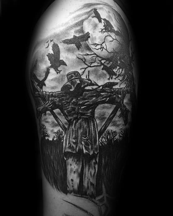 Dark scarecrow tattoo on upper arm with outstretched arms, flying crows, leafless trees, and a full moon in eerie black and grey.