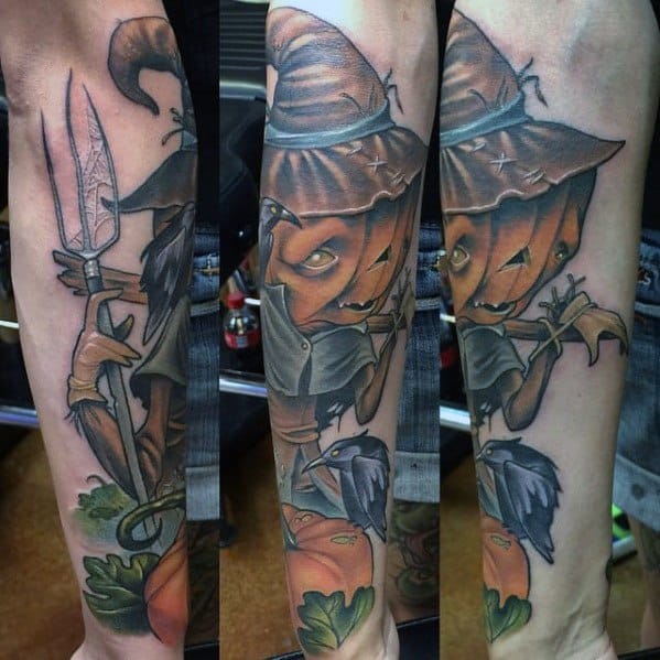Colorful scarecrow tattoo with a sinister pumpkin head, tattered hat, pitchfork, crow, and detailed pumpkins and leaves on the forearm.