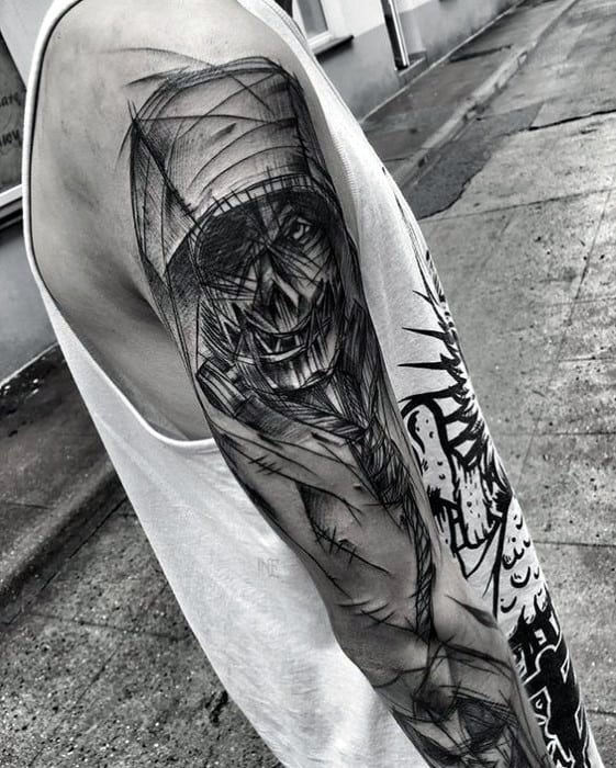 Sketch-style scarecrow tattoo on upper arm with chaotic lines, stitched face, wide grin, and abstract black ink shading.