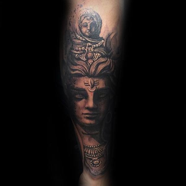 Forearm tattoo of Lord Shiva with a calm, powerful gaze, detailed crown, crescent moon, and ornamental necklace, highlighted with soft shadowing.