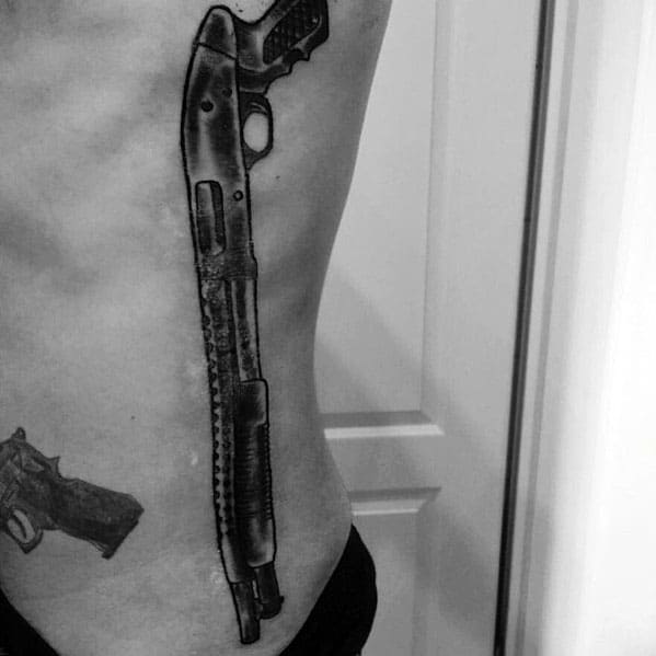 Realistic black and grey shotgun tattoo running along the side of the torso, featuring detailed shading and bold linework for a striking design.