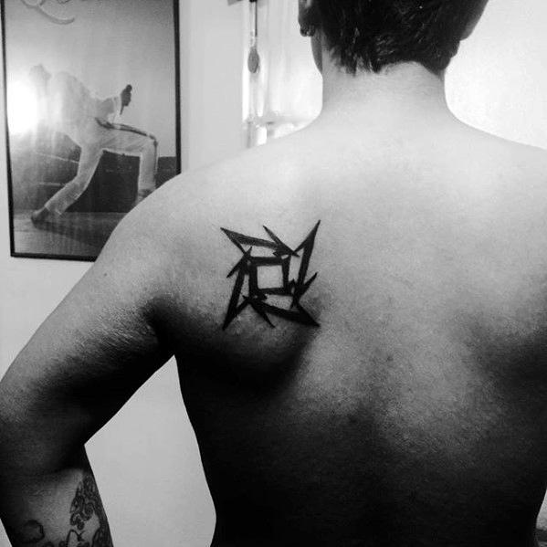 Black Metallica ninja star tattoo on the upper back near the shoulder blade, showcasing a bold and edgy symmetrical design.