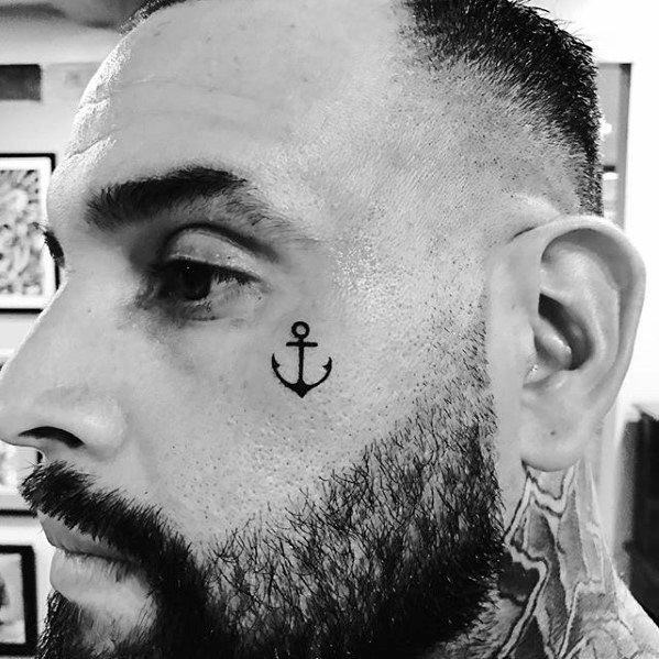 Small black anchor tattoo on the face near the cheekbone, featuring a minimalist design symbolizing strength, stability, and a nautical connection.