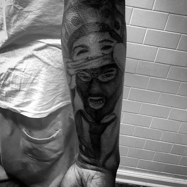 Realistic black and grey forearm tattoo of a person in a ski mask with expressive eyes and mouth, partially covered by hands.