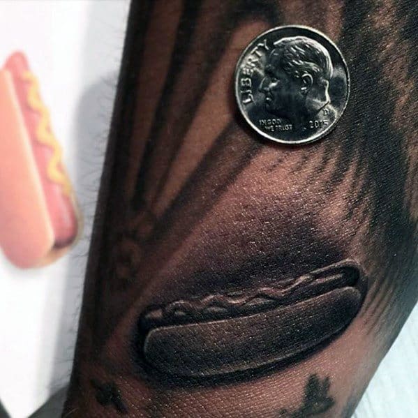 Tattoo of a hyper-realistic hot dog with mustard, appearing three-dimensional on the forearm, with a coin placed for scale.