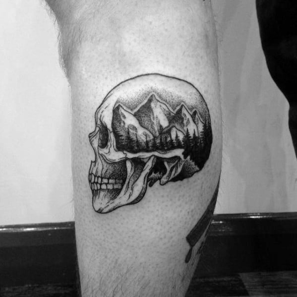 Tattoo of a skull with a mountain and forest scene inside it, on a person's leg