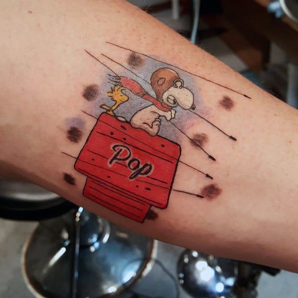 Colorful tattoo of Snoopy as a flying ace with Woodstock on his red doghouse, labeled "Pop," with motion lines and bullet hole effects.