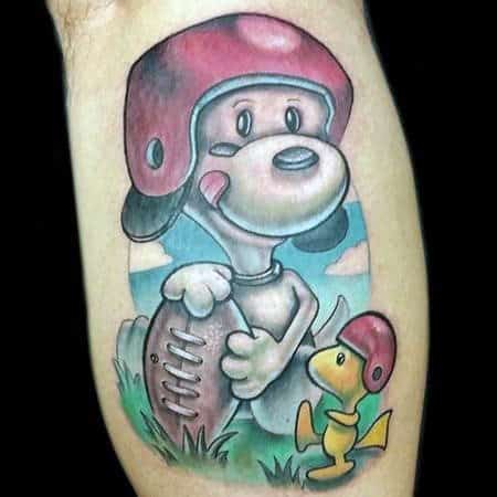 Colorful tattoo of Snoopy and Woodstock wearing red football helmets, with Snoopy holding a football against a blue sky background.