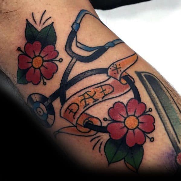 Traditional-style stethoscope tattoo with flowers and a "Dad" banner, honoring a father’s dedication to medicine and healthcare.
