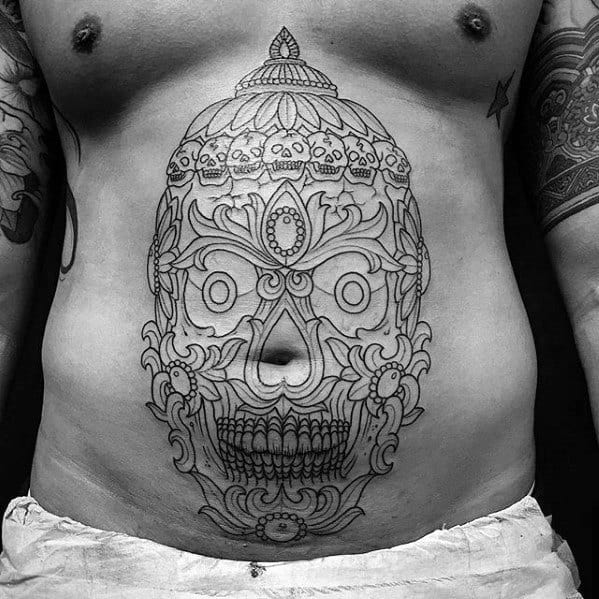 Large Tibetan skull outline tattoo on the stomach with ornamental crown, symmetrical patterns, and fine black linework.