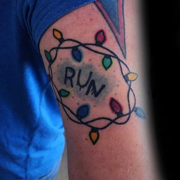 Colorful Stranger Things tattoo of the word “RUN” surrounded by a string of glowing Christmas lights on the upper arm.