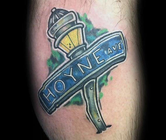 Colorful tattoo of a 'Hoyne Ave' street sign with a vintage-style lamppost, featuring bold blue and yellow tones with a green background.