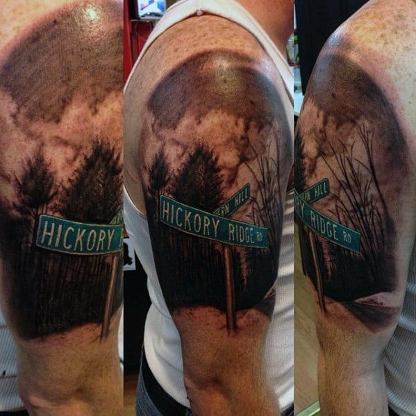 Realistic black and grey tattoo of a 'Hickory Ridge Rd' street sign set against a detailed forest landscape with dark clouds.