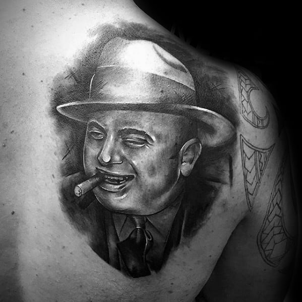 Al Capone tattoo on the shoulder, featuring a detailed black-and-white portrait of the gangster smiling with a cigar and wearing his signature fedora.
