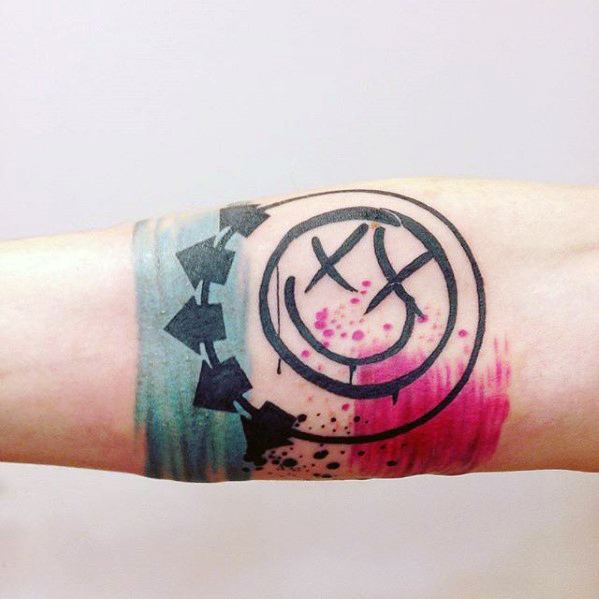 Tattoo of the Blink-182 smiley face logo with black outlines, surrounded by teal and pink watercolor splashes, creating a vibrant and artistic effect on the forearm.