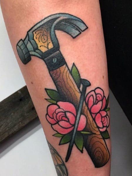 Colorful tattoo of a hammer and nail surrounded by pink roses, symbolizing strength, craftsmanship, and a balance between hard work and beauty.