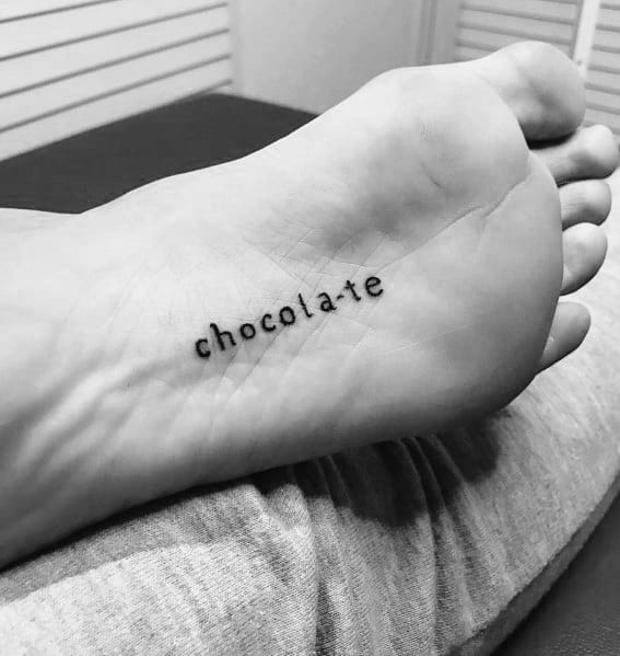 Minimalist tattoo of the word "chocolate" in lowercase letters, placed on the sole of the foot in a simple, clean font.