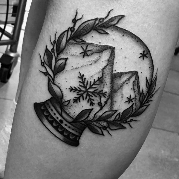 Black and grey Christmas tattoo of a snow globe featuring snowy mountains, a large snowflake, and decorative foliage for an elegant and festive winter design.