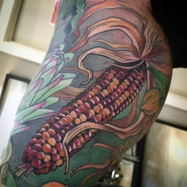 Vibrant tattoo of a multicolored Indian corn cob with detailed husks and surrounding foliage, creating a bold and dynamic design.