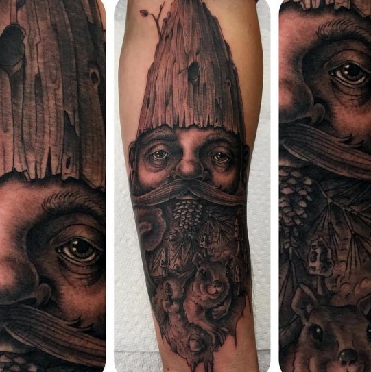 Hyper-realistic black and gray tattoo of a mystical gnome with a wooden-textured hat, intense eyes, and a beard filled with forest animals and foliage.