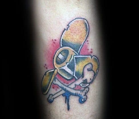 Colorful forearm tattoo of a stylized yellow propeller with crossed bones, featuring bold outlines, shading, and a red splatter background.