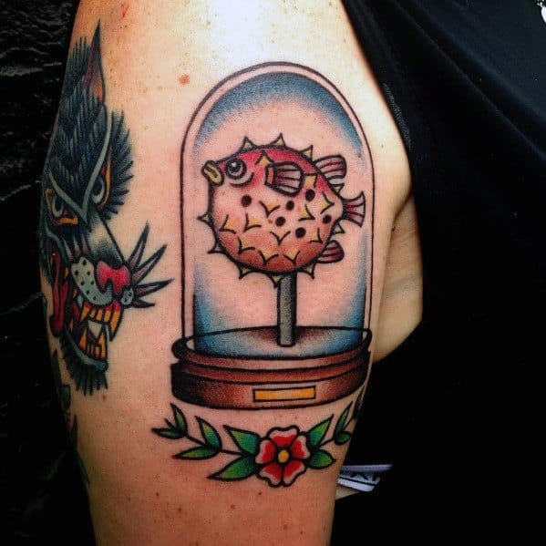 Traditional-style pufferfish tattoo in a glass display case with bold outlines, rich shading, and floral accents for a vintage museum-inspired look.
