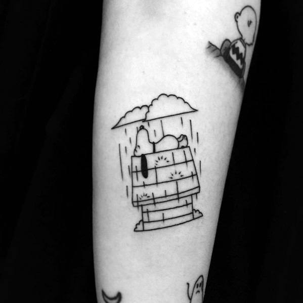 Black and white tattoo of Snoopy lying on his doghouse in the rain, with a small cloud hovering above, placed on a person's forearm.